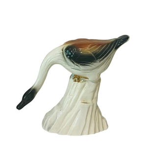 Puede incluir: Una figurita de cerámica blanca de un ganso con marcas marrones, negras y blancas. El ganso está de pie sobre un tocón de árbol de cerámica blanca.