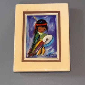 Pode incluir: Uma telha de cerâmica retangular, creme, com uma pintura emoldurada de uma criança nativa americana tocando um tambor. A criança usa uma faixa vermelha e roupas coloridas. O fundo é azul com pinceladas brancas. A assinatura do artista é visível.