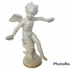Puede incluir: Figurilla de porcelana blanca de un ángel con alas, sosteniendo dos objetos redondos en cada mano. El ángel está de pie sobre una base con un borde dorado.