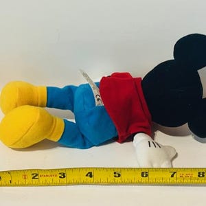 K&ouml;nnte beinhalten: Ein Pl&uuml;sch-Mickey-Mouse-Spielzeug, das ein rotes Hemd und eine blaue Hose tr&auml;gt. Das Spielzeug liegt auf dem R&uuml;cken, die Arme sind ausgestreckt und die Beine sind gebeugt. Das Spielzeug ist etwa 18 cm lang.