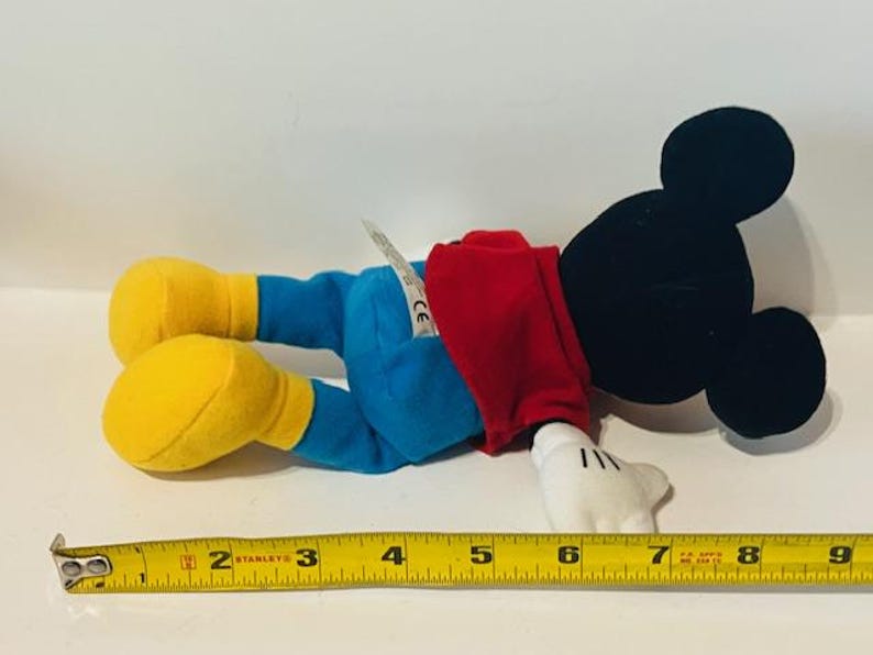 K&ouml;nnte beinhalten: Ein Pl&uuml;sch-Mickey-Mouse-Spielzeug, das ein rotes Hemd und eine blaue Hose tr&auml;gt. Das Spielzeug liegt auf dem R&uuml;cken, die Arme sind ausgestreckt und die Beine sind gebeugt. Das Spielzeug ist etwa 18 cm lang.