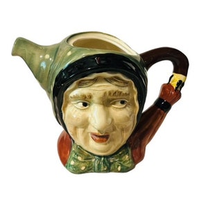 May include: A ceramic teapot shaped like a woman's head with a green and white patterned dress, black hair, and a red and brown handle.