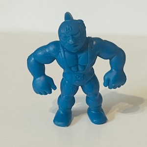 以下が含まれることがあります： 黒いマスクと白黒のストライプの衣装を着たレスラーの青いプラスチック製アクションフィギュア。フィギュアは拳を突き上げており、決意に満ちた表情をしています。