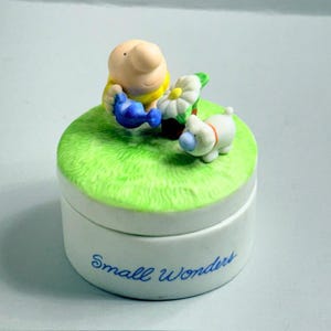 May include: A white ceramic trinket box with a green grassy top. A cartoon figure in a yellow shirt waters a flower with a blue watering can. A white dog with a red collar is also on the top. The words "Small Wonders" are written on the side.