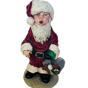 Op de afbeelding: Een klein, rood en wit kerstmanfiguurtje met een witte baard en een groene zak. Hij houdt een bruin speelgoed in zijn andere hand. Het figuurtje staat op een beige voet.