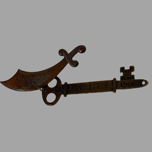 Op de afbeelding: Een vintage souvenir sleutelhanger met een gestileerde sleutel met de woorden "Long Beach 1906" en een decoratief zwaard met het woord "California" erop.