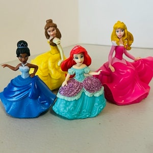 May include: Four Disney princess figurines, each wearing a different coloured gown. The princesses are Belle in yellow, Ariel in teal, Tiana in blue, and Aurora in pink.