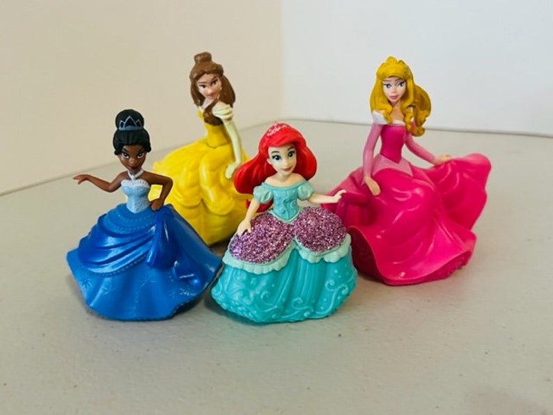 May include: Four Disney princess figurines, each wearing a different coloured gown. The princesses are Belle in yellow, Ariel in teal, Tiana in blue, and Aurora in pink.