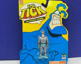 Op de afbeelding: Een plastic action figure van The Tick, een blauwe superheld met een groot hoofd en een insectenachtig gezicht, die een kleinere grijze action figure vasthoudt van een personage met een blauwe riem. De verpakking is geel en blauw met de woorden "The Tick" en "Collectible Figures".