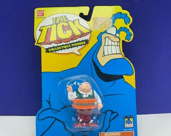 May include: A plastic action figure of The Tick cartoon character, in a blue suit with a red cape, holding a smaller figure of a man with a barrel. The packaging is yellow and blue with the words "The Tick Collectible Figures" on it.