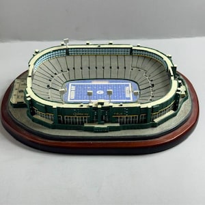 May include: A miniature replica of a football stadium, featuring a green and white design with a blue field. The stadium model is mounted on a dark wooden base, showcasing intricate details of the seating and field markings.
