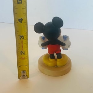 May include: A ceramic figurine of Mickey Mouse holding two blue and white balloons. Mickey is wearing red shorts and yellow shoes. The figurine is standing on a round base.