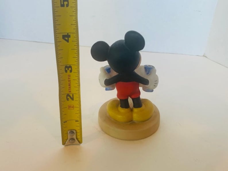 May include: A ceramic figurine of Mickey Mouse holding two blue and white balloons. Mickey is wearing red shorts and yellow shoes. The figurine is standing on a round base.