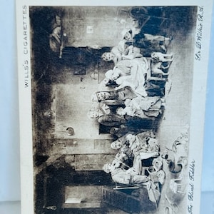 May include: Black and white vintage postcard featuring a group of people gathered around a blind fiddler. The text on the postcard reads "The Blind Fiddler" and "Wills's Cigarettes".
