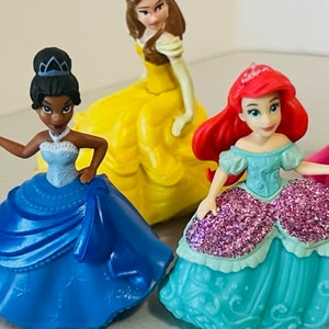 May include: Four Disney princess figurines wearing ball gowns. The princesses are Belle, Tiana, Ariel, and Snow White. The figurines are made of plastic and are about 7.5 cm tall.