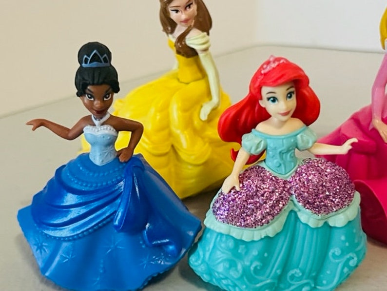 May include: Four Disney princess figurines wearing ball gowns. The princesses are Belle, Tiana, Ariel, and Snow White. The figurines are made of plastic and are about 7.5 cm tall.