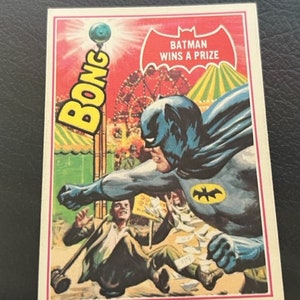Könnte beinhalten: Eine Vintage-Batman-Sammelkarte mit einer farbenfrohen Illustration von Batman, der einen Preis auf einem Jahrmarkt gewinnt. Die Karte trägt den Text "Batman Wins A Prize" und "BONG" in großen Buchstaben.