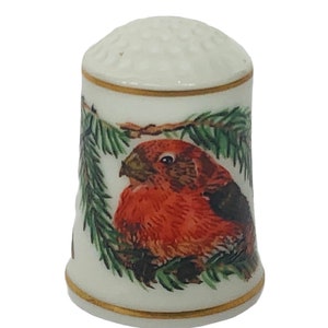 Op de afbeelding: Een witte porseleinen vingerhoed met een gouden rand en een geschilderd beeld van een rode vogel die op een groene dennentak zit.