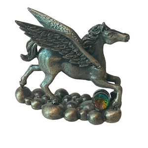 May include: A silver Pegasus figurine with wings outstretched, standing on a cloud-like base with a colorful glass orb. The figurine is detailed with textured wings and a flowing mane.