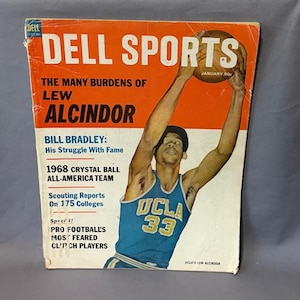 Może przedstawiać: Okładka magazynu Dell Sports z Lewem Alcindorem w koszulce UCLA Bruins. Okładka jest pomarańczowo-biała, z tekstem "The Many Burdens of Lew Alcindor" i "January 50¢". Kolekcjonerski magazyn sportowy.