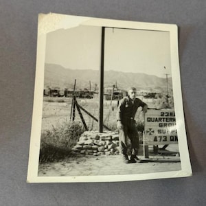 May include: A monochrome photograph showing a man in uniform beside a sign. The sign reads "23rd Quartermaster Group" and "473 QM". The background features a field, mountains, and military vehicles.