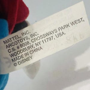 K&ouml;nnte beinhalten: Ein wei&szlig;es Etikett mit schwarzem Text, der "MATTEL, INC. ARCOTOYS, INC. C.S. # 9008, CROSSWAYS PARK WEST, WOODBURY, NY 11797, USA. MADE IN CHINA &copy;DISNEY" lautet.