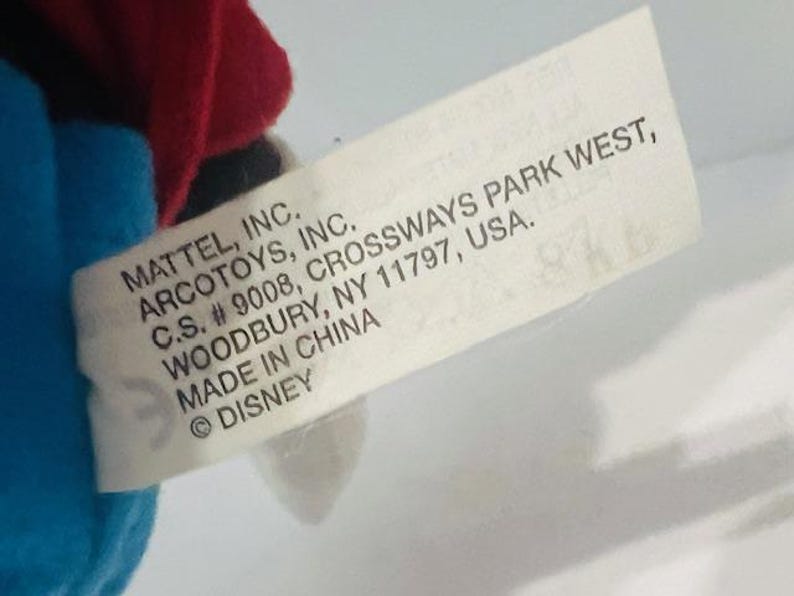 K&ouml;nnte beinhalten: Ein wei&szlig;es Etikett mit schwarzem Text, der "MATTEL, INC. ARCOTOYS, INC. C.S. # 9008, CROSSWAYS PARK WEST, WOODBURY, NY 11797, USA. MADE IN CHINA &copy;DISNEY" lautet.