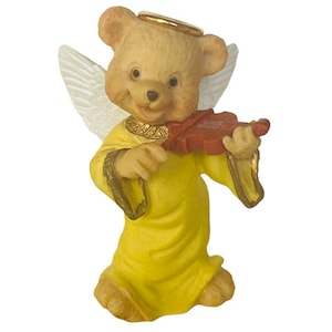 Pode incluir: Uma pequena figura de cerâmica de um anjo urso de peluche tocando violino. O urso está vestindo uma túnica amarela e tem uma auréola acima de sua cabeça. O violino é vermelho.