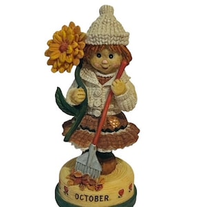 May include: A figurine of a girl wearing a white knit hat and a brown dress holding a broom and a yellow flower. The base of the figurine is decorated with fall leaves and the word "OCTOBER".