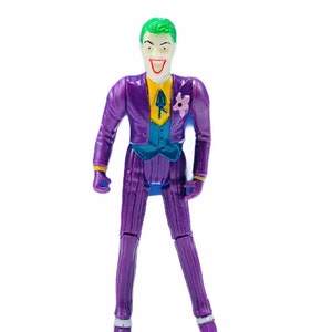 Può includere: Una action figure vintage del Joker, un supercriminale della DC Comics. La figura indossa un abito a righe viola con un gilet verde acqua, camicia gialla e un fiore rosa sull'occhiello. Il Joker ha i capelli verdi e un ampio sorriso sinistro.