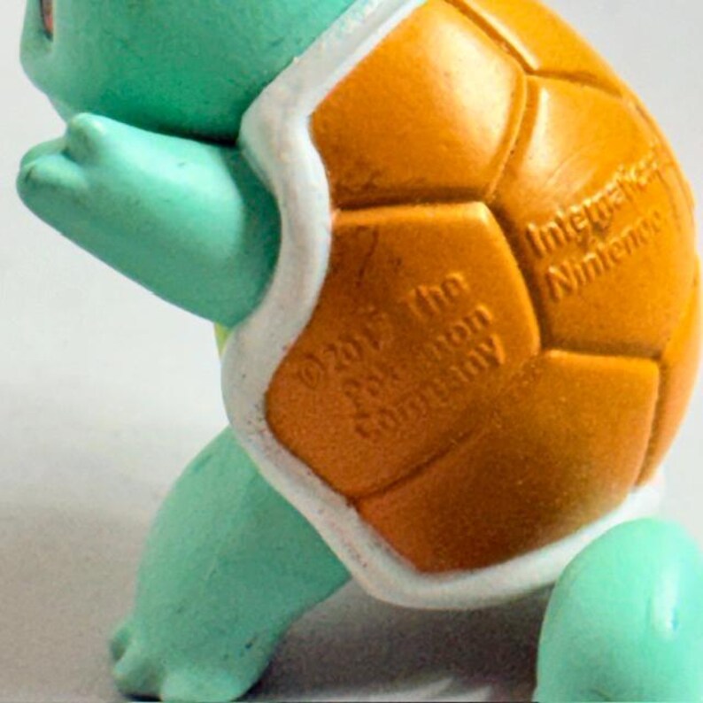 Puede incluir: Primer plano de una figura de Squirtle de color turquesa. El caparaz&oacute;n naranja tiene un patr&oacute;n texturizado y se puede ver el texto "International Nintendo" y "&copy;2017 The Pokemon Company".