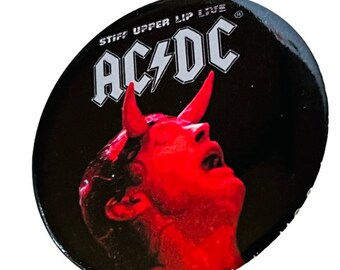 May include: A black and white button with a red devil head and the text "AC/DC" in white with a lightning bolt. The text "Stiff Upper Lip Live" is also in white around the top of the button.