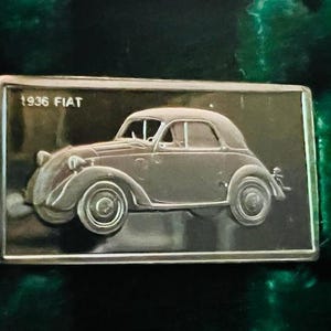 Op de afbeelding: Een zilverkleurige metalen plaquette met de tekst "1936 FIAT" en een reliëfbeeld van een oldtimer.