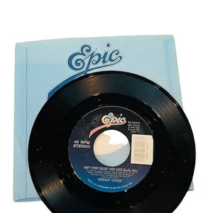 Puede incluir: Un disco de vinilo negro de 45 RPM con una etiqueta blanca. La etiqueta tiene el texto "Epic", "34-72464", "205 724644", "Can't Stop Fallin' Into Love (Radio Mix)", "Cheap Trick", y "45 RPM Stereo".