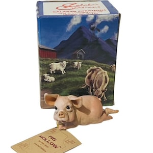 May include: A small, pink pig figurine with a tag that reads "Pig Hollow" and a box with a rural scene and the text "Calabar Creations".