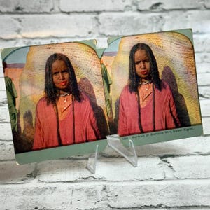 Puede incluir: Una tarjeta estereoscópica vintage con una fotografía coloreada de una persona de cabello oscuro que viste una prenda roja y un collar. El texto en la parte inferior derecha dice 1380. Portrait of Bisharin Girl, Upper Egypt. Tarjeta en soporte.