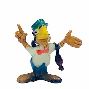 May include: A yellow and brown cartoon bird figurine wearing a green hat and a red bow tie. The bird is holding its right wing up and its left arm out to the side. The figurine is made of plastic.