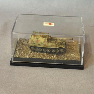 May include: A model tank in a clear display case. The tank is painted in camouflage colors of brown, green, and tan. The display case sits on a black base and includes a small label that reads "CAN DO".