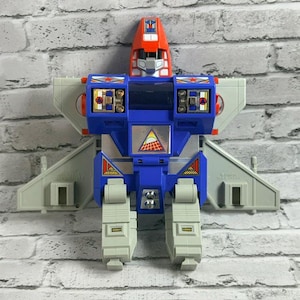 May include: A vintage plastic robot action figure featuring a red head, blue torso, and light gray wings and legs. The chest displays a triangular sticker with a sun and grid graphic, while the shoulders feature red star decals and metallic details.