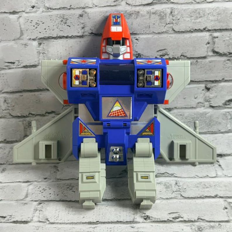 May include: A vintage plastic robot action figure featuring a red head, blue torso, and light gray wings and legs. The chest displays a triangular sticker with a sun and grid graphic, while the shoulders feature red star decals and metallic details.