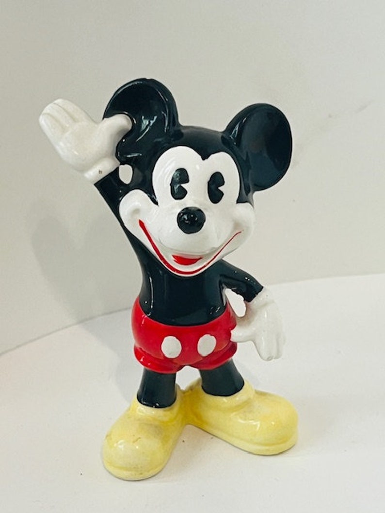 May include: A black and white ceramic figurine of Mickey Mouse wearing red shorts with white polka dots. He is waving with his right hand and his left hand is on his hip. He has a big smile on his face.