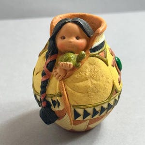 May include: A small, round figurine of a Native American child holding a green turtle. The child is inside a yellow and orange container with geometric patterns and a green gem. The child has black hair in a braid.