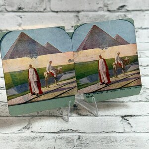Puede incluir: Una tarjeta estereoscópica vintage con una escena coloreada de las pirámides de Gizeh en Egipto. La imagen muestra dos figuras con túnicas blancas y capas rojas, una caminando y otra montada en un burro, con el texto The Pyramids at Ghizeh from the Tramway, Egypt.
