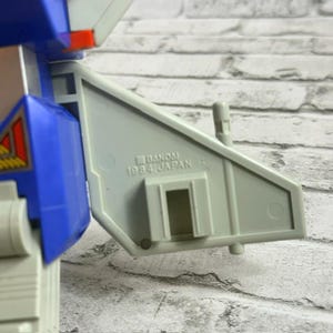 May include: A vintage plastic robot toy component in light gray and blue, featuring a molded stamp that reads BANDAI 1984 JAPAN. The piece has a geometric wing-like structure with a rectangular cutout and a small vertical post on the top edge.