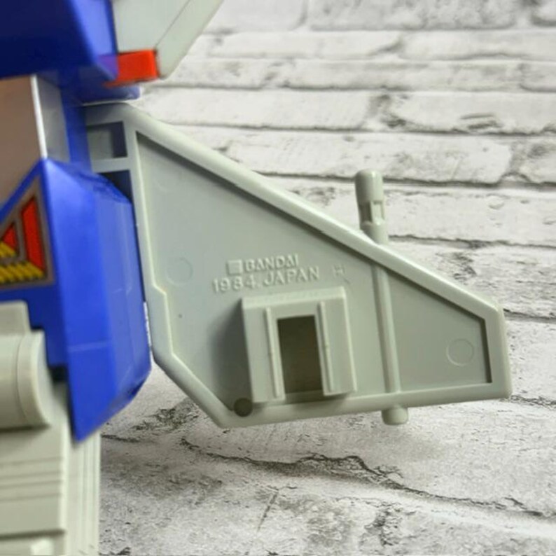 May include: A vintage plastic robot toy component in light gray and blue, featuring a molded stamp that reads BANDAI 1984 JAPAN. The piece has a geometric wing-like structure with a rectangular cutout and a small vertical post on the top edge.