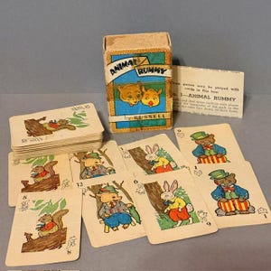 May include: Vintage "Animal Rummy" card game by Russell. The box features a blue and gold design with two bear-like characters. The cards depict various animals in colorful outfits. A small instruction card is included.