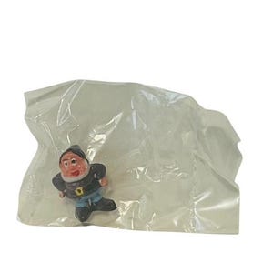 May include: A small plastic figurine of a dwarf wearing a blue and black outfit. The dwarf has a red and white painted face and is packaged in a clear plastic bag.