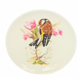 Puede incluir: Un plato de cerámica blanco con un diseño pintado a mano de un jilguero posado en una planta de cardo con flores rosas. El pájaro tiene rayas negras y blancas en sus alas y una cabeza roja.