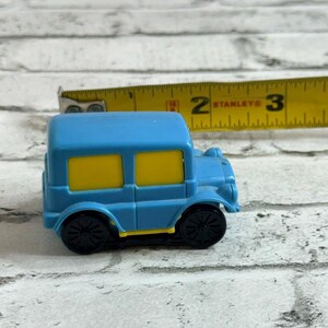 May include: A small blue toy car with yellow windows and black wheels, positioned next to a yellow Stanley measuring tape that shows the vehicle is approximately 6.35 cm in length. The toy car features a boxy design with a bright blue body.
