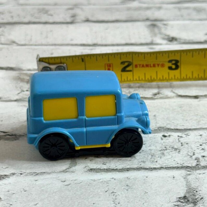 May include: A small blue toy car with yellow windows and black wheels, positioned next to a yellow Stanley measuring tape that shows the vehicle is approximately 6.35 cm in length. The toy car features a boxy design with a bright blue body.